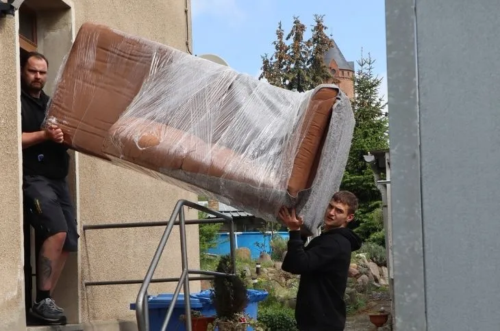 zwei umzugshelfer tragen eingewickeltes sofa aus wohngebaeude